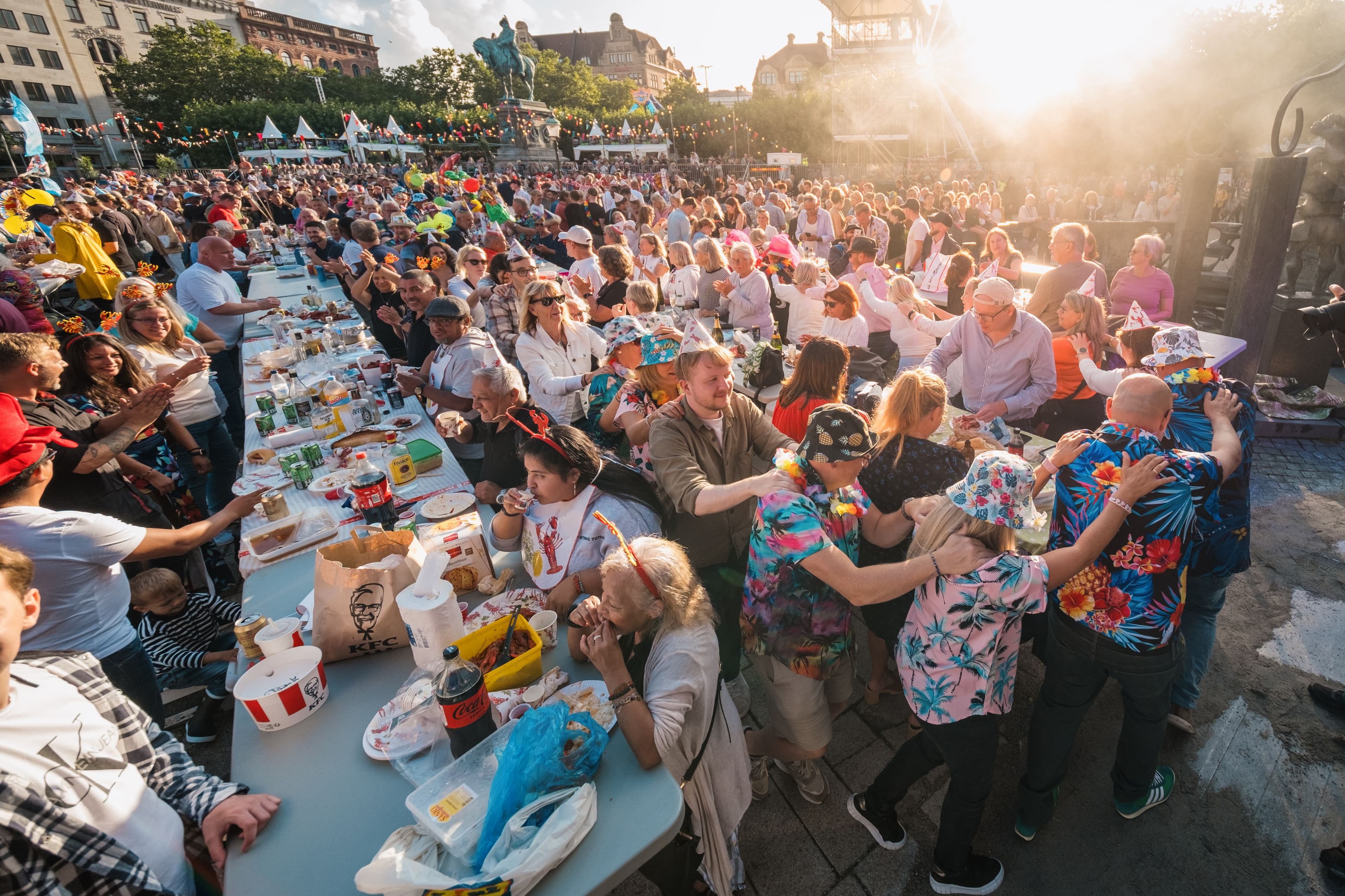 Festklädda människor äter kräftor och firar tillsammans vid långbord på ett soligt torg under Malmöfestivalen.