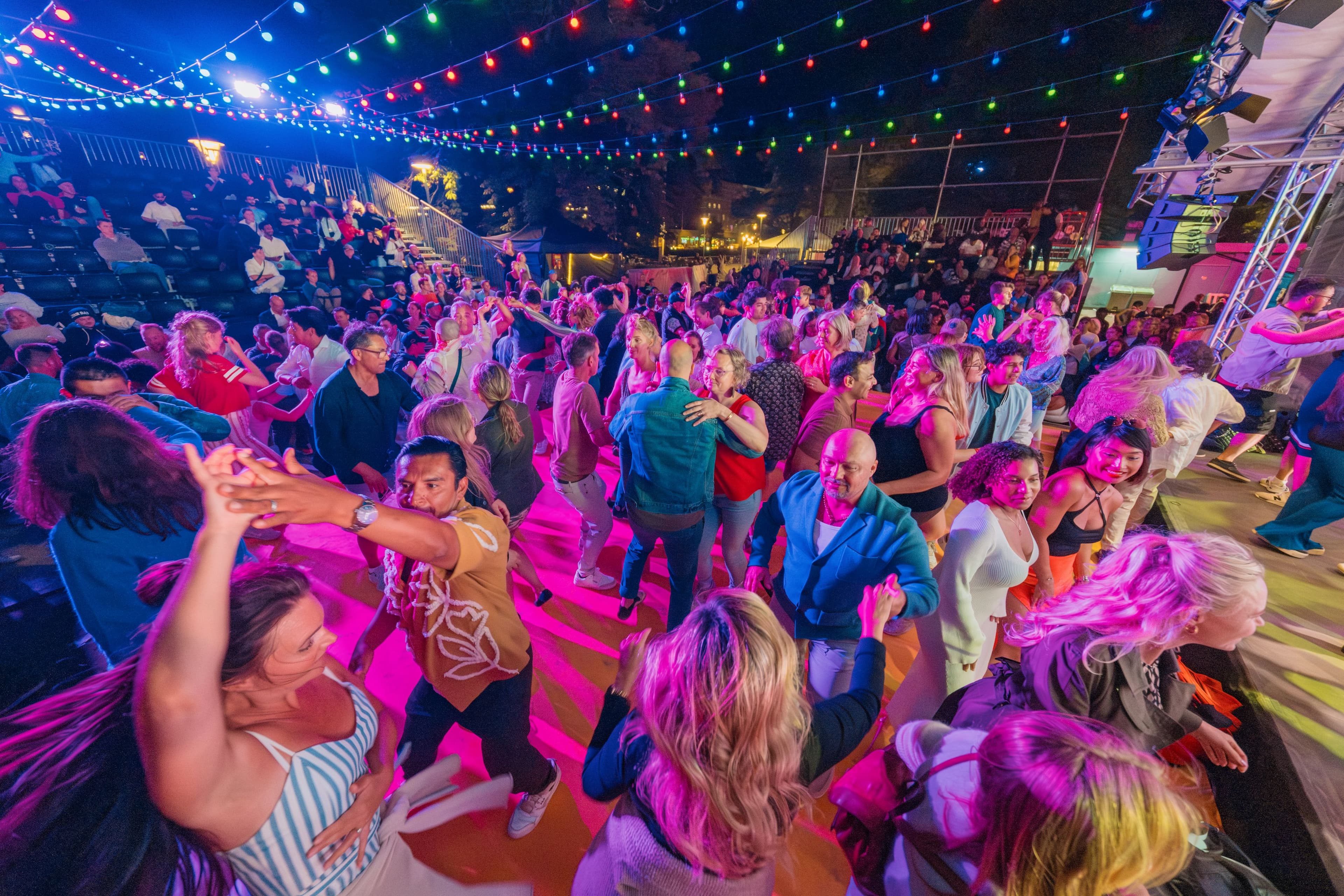 Människor dansar under färgglad belysning på en utescen med publik i bakgrunden.