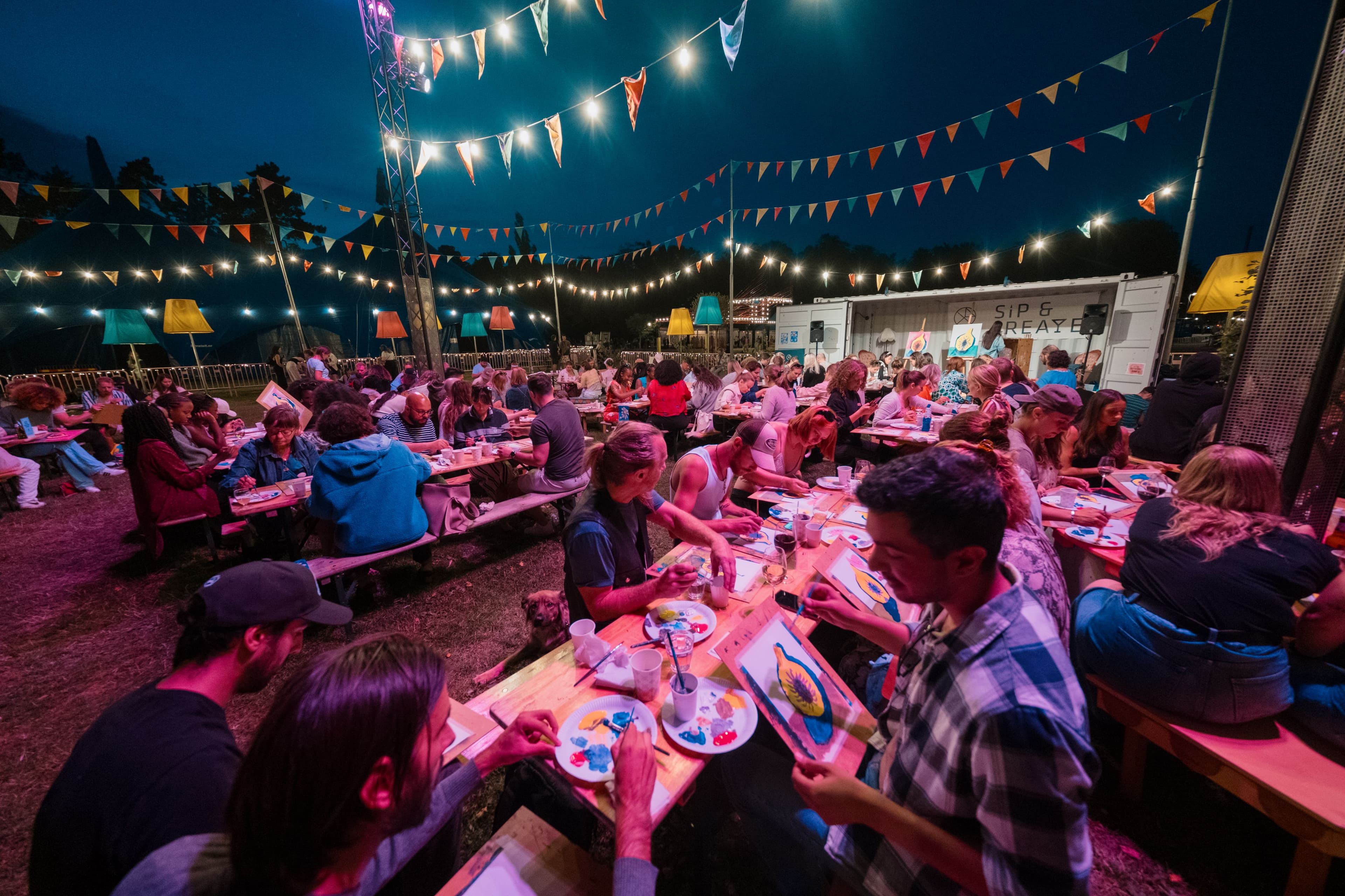 Personer sitter vid långbord och målar tavlor under kvällen på en utomhusworkshop på Malmöfestivalen.
